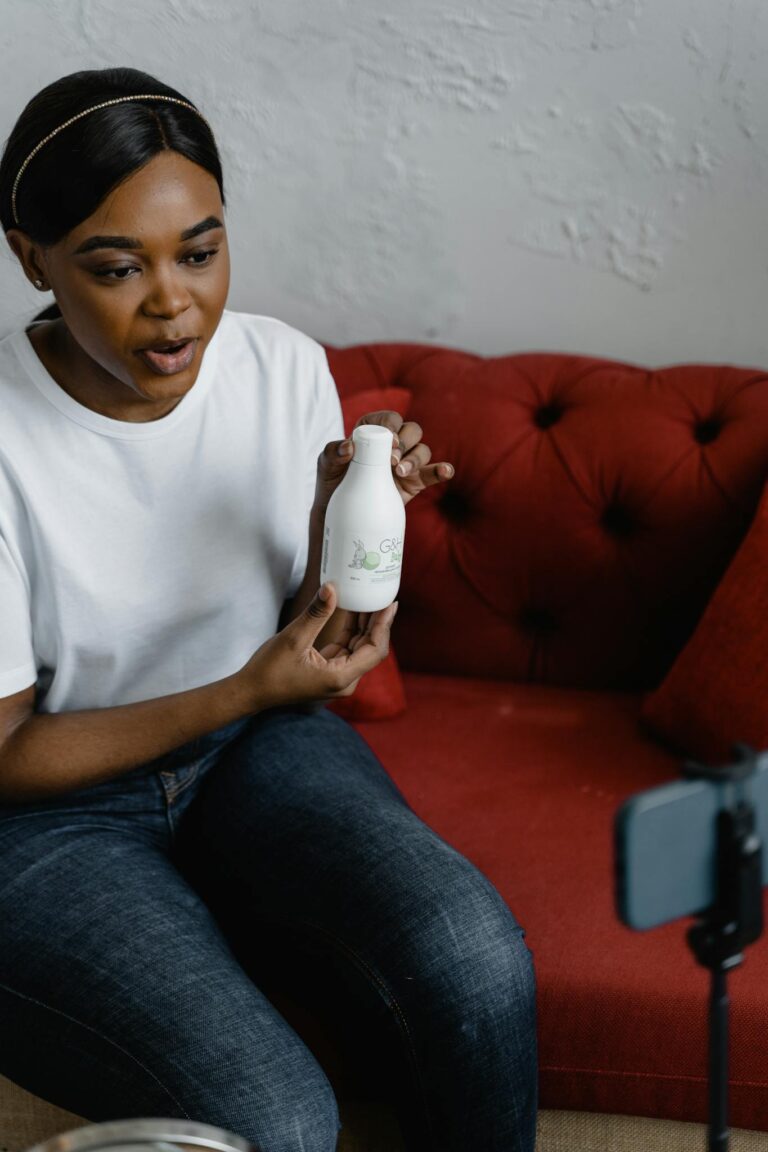African American woman presenting a cosmetic product while vlogging on a red sofa.